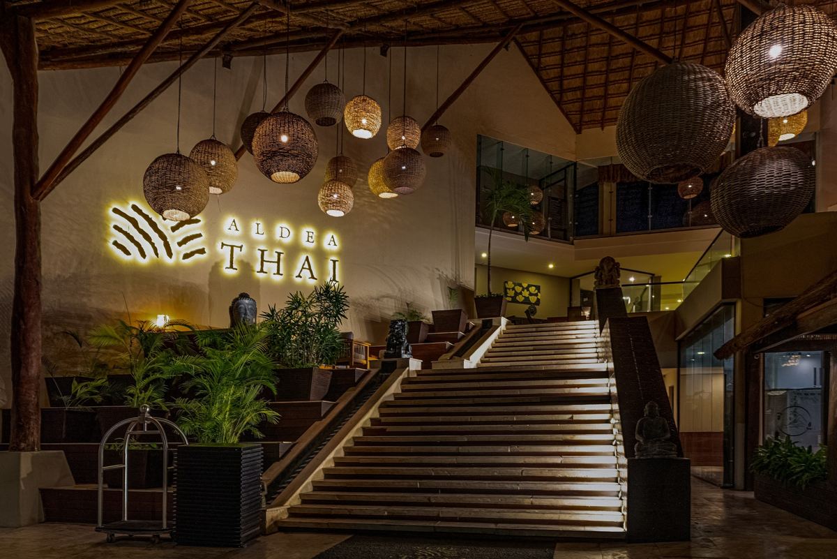 La entrada de Aldea Thai de noche: el letrero dorado iluminado, las lámparas de ratán y la escalera de piedra